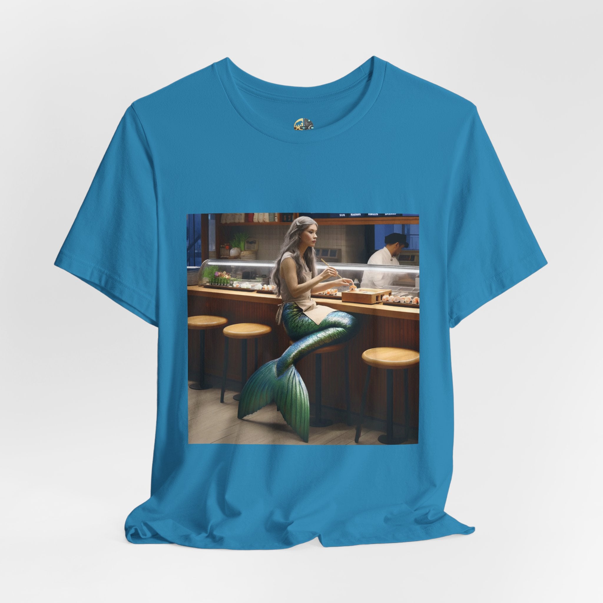 Aqua blue T-shirt featuring a graphic of a mermaid wearing a chef’s apron sitting at a sushi bar and eating sushi, with a chef behind the counter