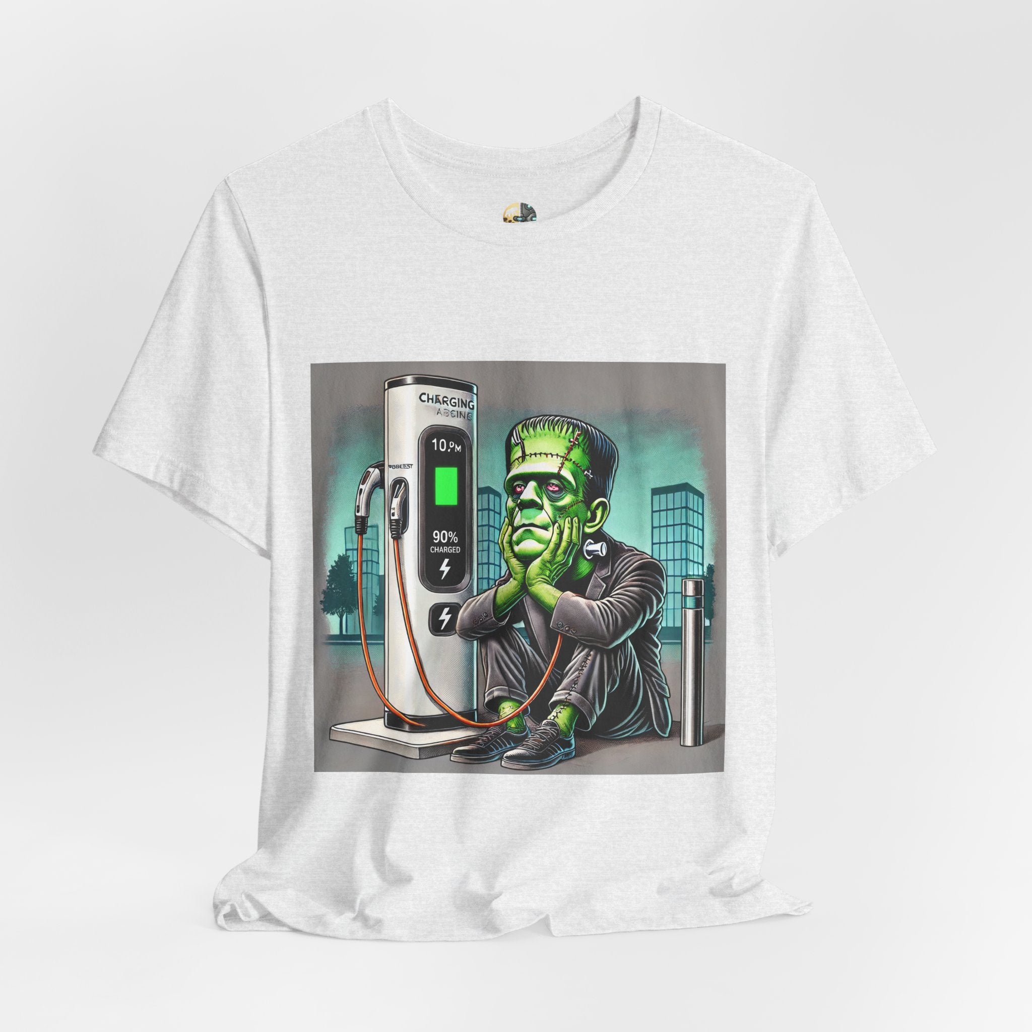 Ash color T-shirt featuring a graphic of Frankenstein’s monster sitting at an electric charging station, plugged in and waiting for the charge to complete, with office buildings in the background