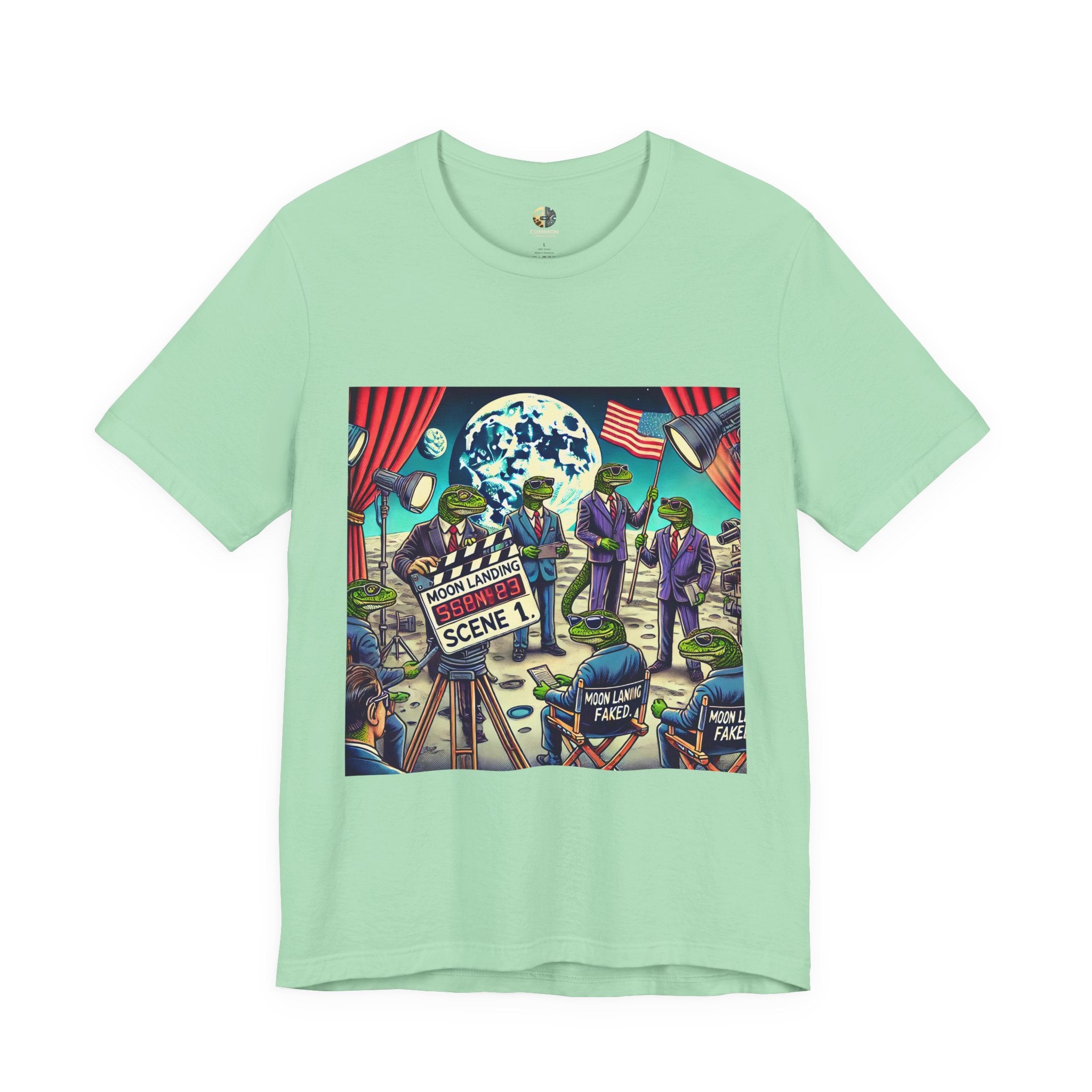 Mint green T-shirt featuring a graphic of lizard people in suits on a movie set staging a moon landing scene, with studio lights and a clapperboard