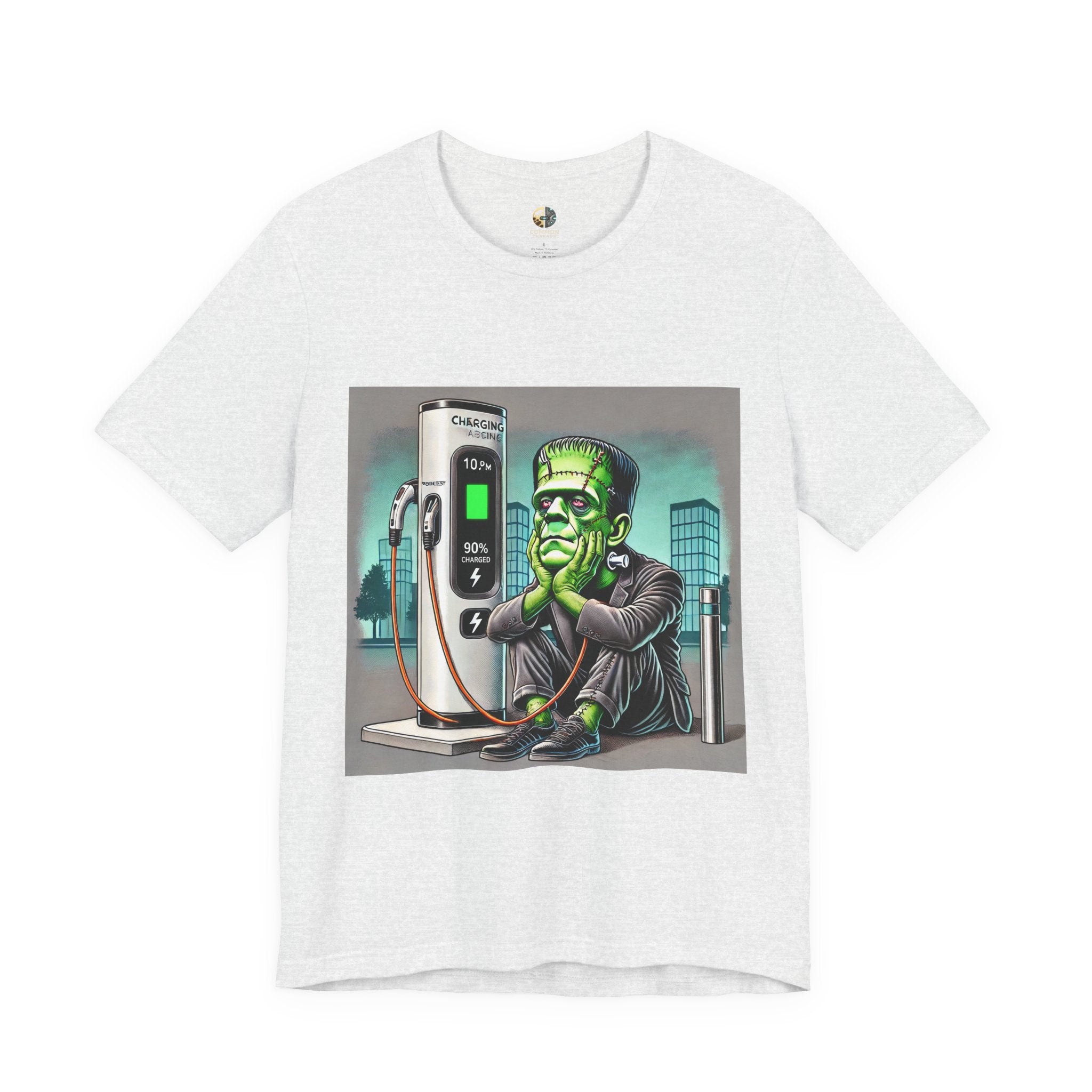Ash color T-shirt featuring a graphic of Frankenstein’s monster sitting at an electric charging station, plugged in and waiting for the charge to complete, with office buildings in the background