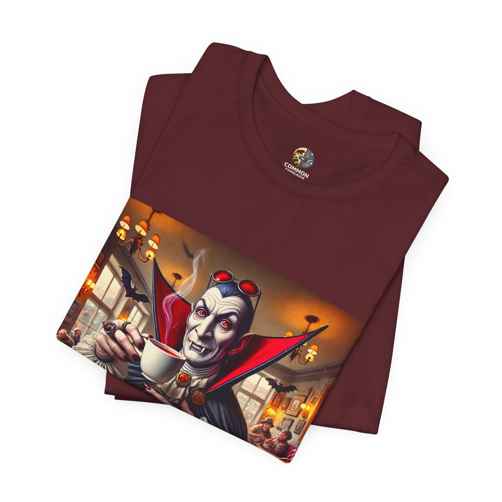 Folded maroon color T-shirt featuring a classic vampire dressed like Count Dracula sitting in a café, wearing red sunglasses and drinking from a teacup filled with red liquid