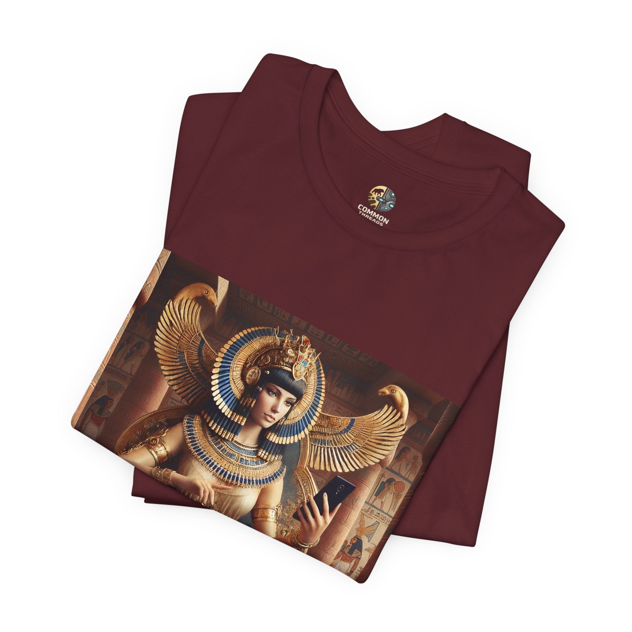 Folded maroon color T-shirt featuring a graphic of Cleopatra wearing an ornate headdress and gold jewelry, seated on a throne and holding a smartphone for a selfie, with temple architecture in the background