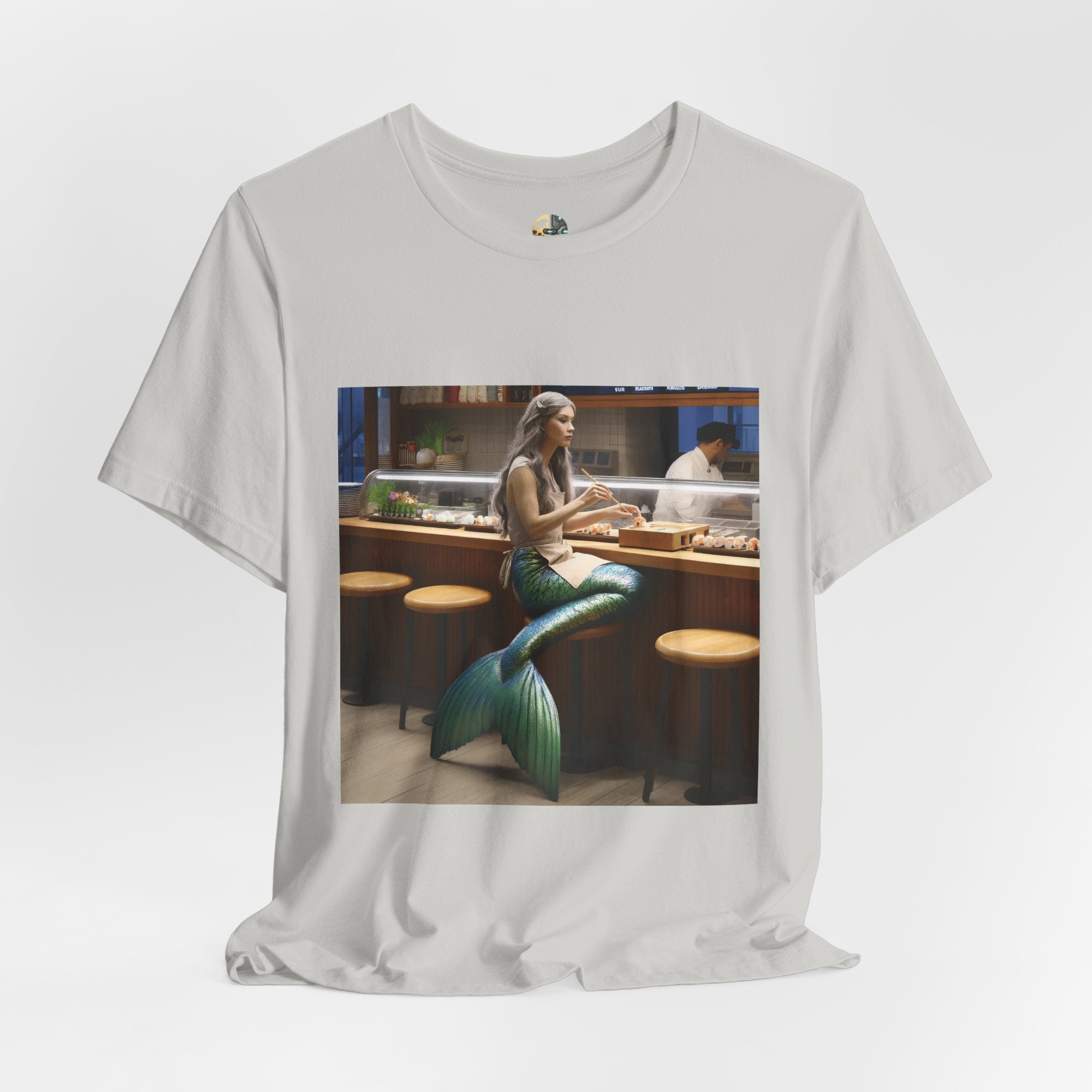 Silver T-shirt featuring a graphic of a mermaid wearing a chef’s apron sitting at a sushi bar and eating sushi, with a chef behind the counter