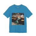 Aqua blue T-shirt featuring a graphic of a mermaid wearing a chef’s apron sitting at a sushi bar and eating sushi, with a chef behind the counter
