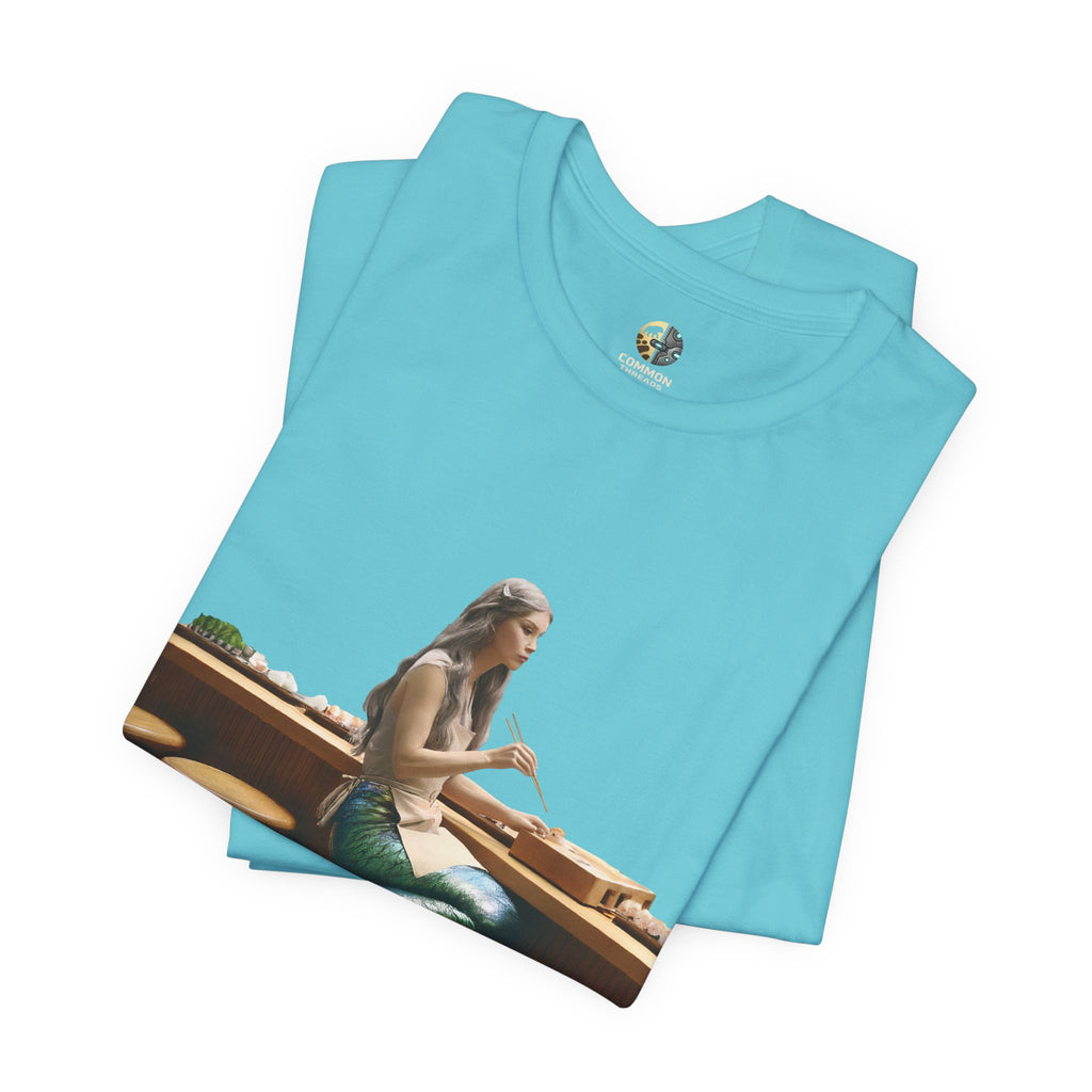 Folded turquoise T-shirt with graphic of a mermaid wearing a chef’s apron sitting at a sushi bar and eating sushi with chopsticks