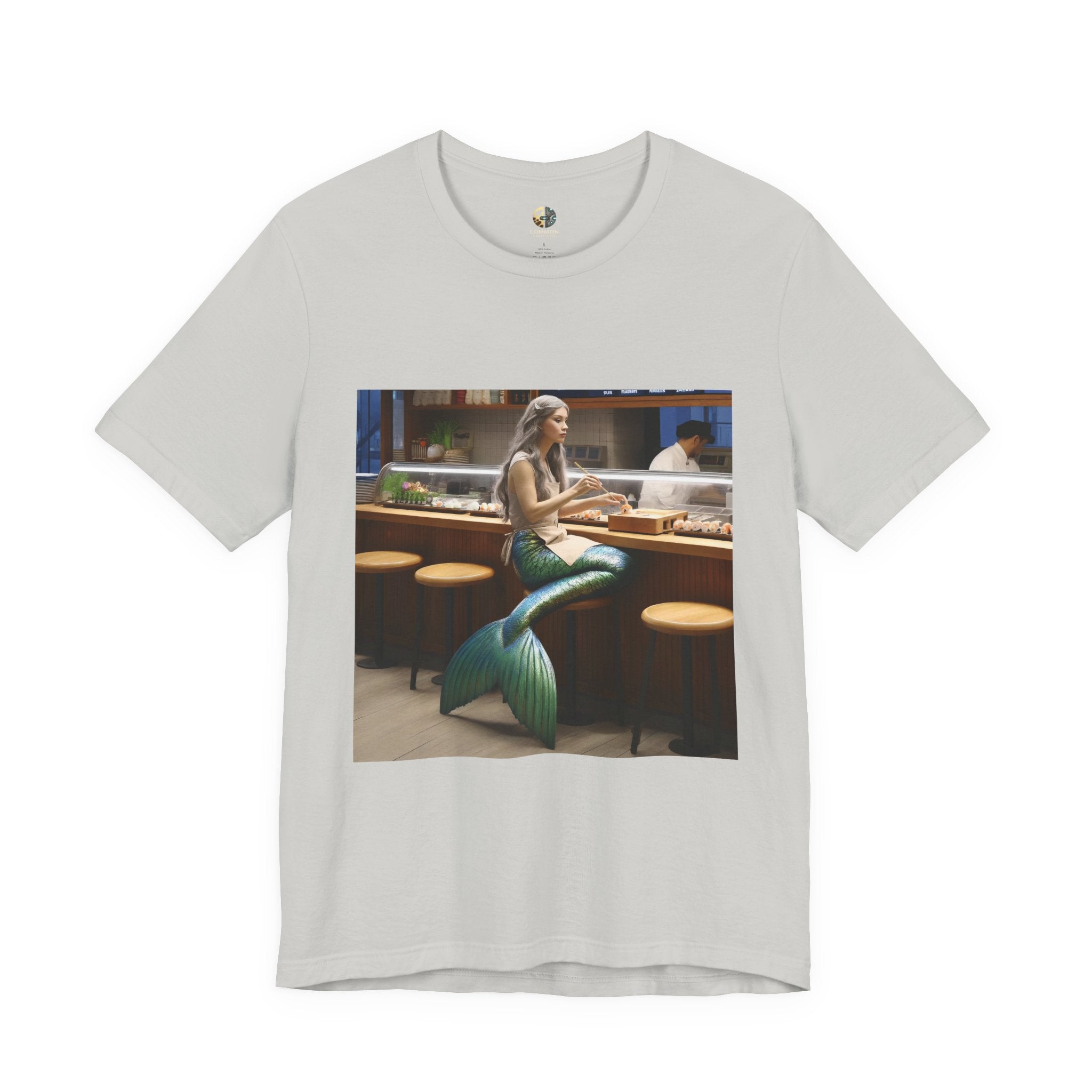 Silver T-shirt featuring a graphic of a mermaid wearing a chef’s apron sitting at a sushi bar and eating sushi, with a chef behind the counter