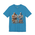 Aqua blue T-shirt featuring a graphic of a caveman in animal pelts standing beside a chrome-plated astronaut, with the text ‘Common Threads‘